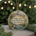 A round ornament with a scenic landscape of mountains, trees, and a river, featuring a duck swimming in the water. The ornament is hanging from a string and is placed on a surface with pine branches.