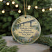 A round ornament with a scenic image of a bridge and a river, surrounded by greenery, is displayed on a surface with Christmas lights in the background.