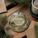 A round ornament with a scenic image of a cabin and a waterfall, surrounded by pine branches and a cup of coffee on a wooden surface.