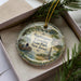 A decorative ornament in a box, featuring a scenic landscape with a boat, a monument, and people enjoying the outdoors.