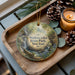 A wooden tray with a decorative ornament featuring a scenic waterfall and greenery, accompanied by pine cones and a lit candle.