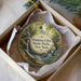 A circular ornament with a painting of a waterfall and greenery, placed in a box with a white tissue paper lining.
