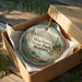 A round ornament in a wooden box, featuring a scenic landscape with mountains, trees, and a cabin, with the text "Wayfarers State Park, Montana" printed on it.
