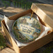A circular ornament in a box, featuring a scenic landscape with a castle, trees, and water.