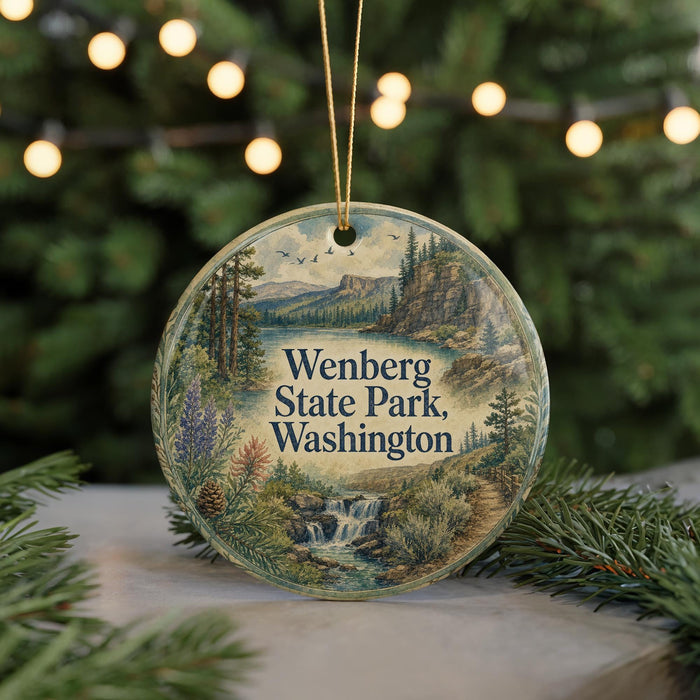 A circular ornament with a scenic landscape of Wenberg State Park, Washington, featuring a waterfall and pine trees, is displayed on a table with Christmas lights in the background.