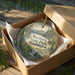 A circular ornament with a scenic landscape of Wenberg State Park in Washington, featuring mountains, waterfalls, and pine trees. The ornament is displayed in a cardboard box with a gold string.