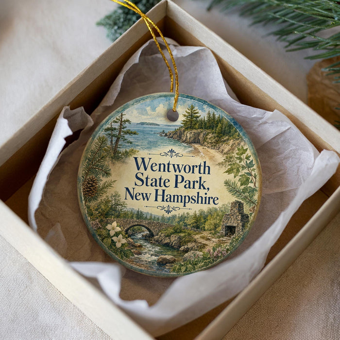 A decorative ornament in a box, featuring a scenic landscape with a river, bridge, and pine trees.