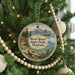 A round ornament hanging on a Christmas tree, featuring a scenic landscape with mountains, a lake, and a sailboat.