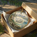 A decorative ornament in a wooden box, featuring a scenic landscape with mountains, trees, and a lake, along with the text "West Shore State Park, Montana".