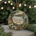 A decorative ornament featuring a deer standing in front of a scenic landscape, with a body of water and a waterfall in the background. The ornament is hanging from a string and is placed on a surface with pine branches.