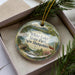 A decorative ornament in a box, featuring a scenic landscape with a lake, mountains, and pine trees.