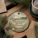A decorative ornament with a scenic landscape of Whitefish Dunes State Park, Wisconsin, featuring pine trees, a lake, and a bird in flight. The ornament is hanging on a wooden surface, surrounded by pine branches and a cup of coffee.