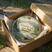 A round ornament in a wooden box, featuring a scenic landscape with mountains, a lake, and a canoe, with the text "Whitefish Lake State Park, Montana" printed on it.