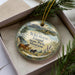 A decorative ornament with a scenic landscape featuring wild horses, pine trees, and mountains, placed in a box with a pine branch.