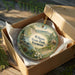 A decorative ornament in a box, featuring a scenic landscape with mountains, pine trees, and a cabin, with the text "Wilgus State Park, Vermont" printed on it.