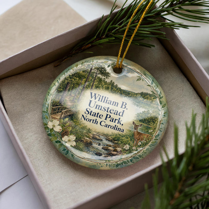 A decorative ornament in a box, featuring a scenic landscape with a deer and trees, with the text "William B. Umsstead State Park North Carolina" printed on it.