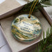 A decorative ornament in a box, featuring a scenic landscape with a windmill, mountains, and a river.