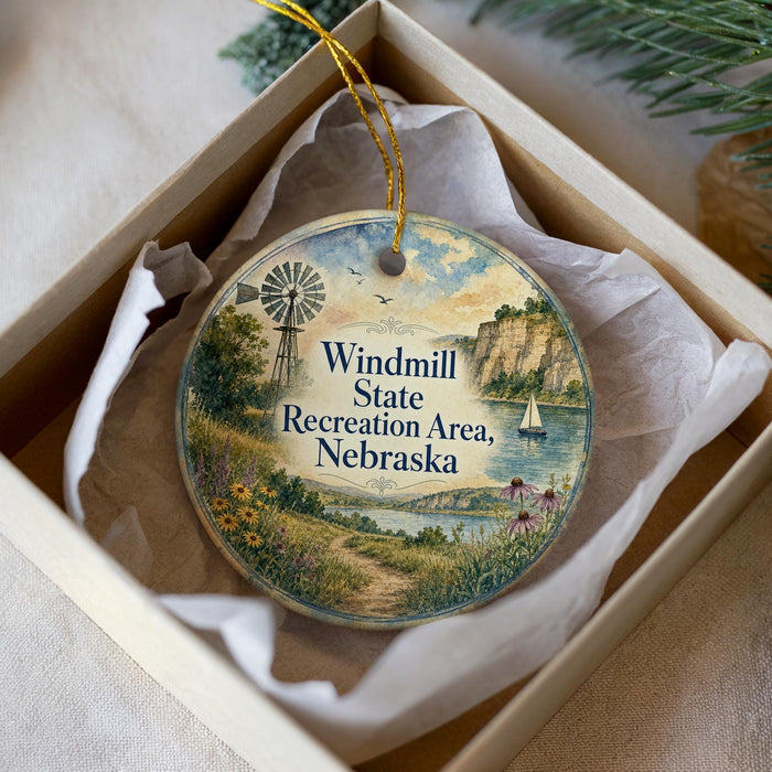 A decorative ornament in a white box, featuring a scenic landscape with a windmill, a sailboat, and a path leading to a lake.