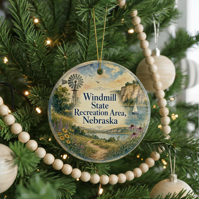 A round ornament hanging on a Christmas tree, featuring a scenic landscape with a windmill, a path, and a body of water.