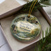 A decorative ornament with a scenic image of Woodford State Park, Vermont, displayed in a box with pine branches.