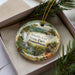 A decorative ornament in a box, featuring a scenic landscape with a waterfall and a cabin, with the text "Worlds End State Park, Pennsylvania" printed on it.