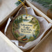 A decorative ornament in a white box, featuring a scenic landscape with a lake, a small building, and a bird.