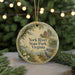 A round ornament with a painting of a house and a bird, surrounded by greenery, hangs in front of a blurred Christmas tree with lights.