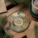 A decorative ornament featuring a scenic landscape with a waterfall, mountains, and a cabin, hanging on a wooden surface.