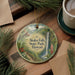 A circular ornament with a scenic image of Akaka Falls State Park in Hawaii, featuring a waterfall, lush greenery, and pine trees.