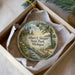 A round ornament with a lighthouse and a scenic landscape, placed in a box with a white tissue paper lining.
