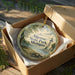 A decorative ornament in a wooden box, featuring a scenic landscape with a lighthouse and trees, with the text "Algonac State Park, Michigan" printed on it.