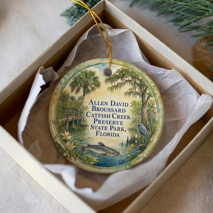 A round ornament with a painting of a catfish and a bird, surrounded by greenery, placed in a box with a white tissue paper lining.