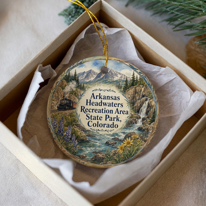 A decorative ornament in a box, featuring a scenic landscape with mountains, a river, and a train.