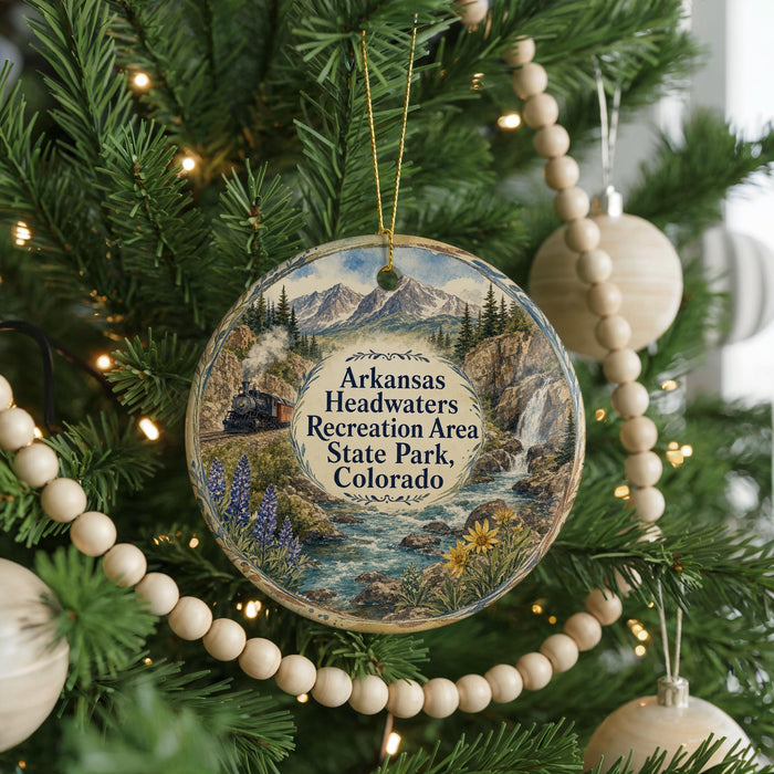 A Christmas ornament hanging on a tree, featuring a scenic landscape with mountains, a river, and a train.