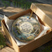 A decorative ornament in a cardboard box, featuring a scenic landscape with mountains, water, and greenery.