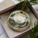 A decorative ornament in a box, featuring a scenic landscape with a waterfall and pine trees.