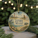 A round ornament with a lighthouse and a horse, surrounded by greenery and a beach scene, hangs in front of a Christmas tree with twinkling lights.