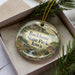 A decorative ornament in a box, featuring a scenic landscape with a river, buildings, and greenery.