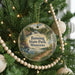 A circular ornament hanging on a Christmas tree, featuring a scenic landscape with a river, pine trees, and a waterfall.