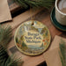 A round ornament with a scenic landscape of Baraga State Park, Michigan, featuring a waterfall and pine trees, is displayed on a wooden surface alongside a cup of coffee and some papers.