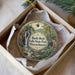 A circular ornament with a scenic image of a waterfall and forest, placed in a box with a white tissue paper lining.