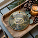 A decorative ornament featuring a scenic image of Baxter State Park, Maine, with a moose and a mountain in the background. The ornament is displayed on a wooden tray, accompanied by pine cones and a lit candle.