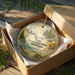 A round ornament in a brown box, featuring a scenic landscape with a lighthouse and a body of water.