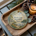 A decorative ornament with a scenic lake and park scene, featuring a bird, pine cones, and a candle on a wooden tray.