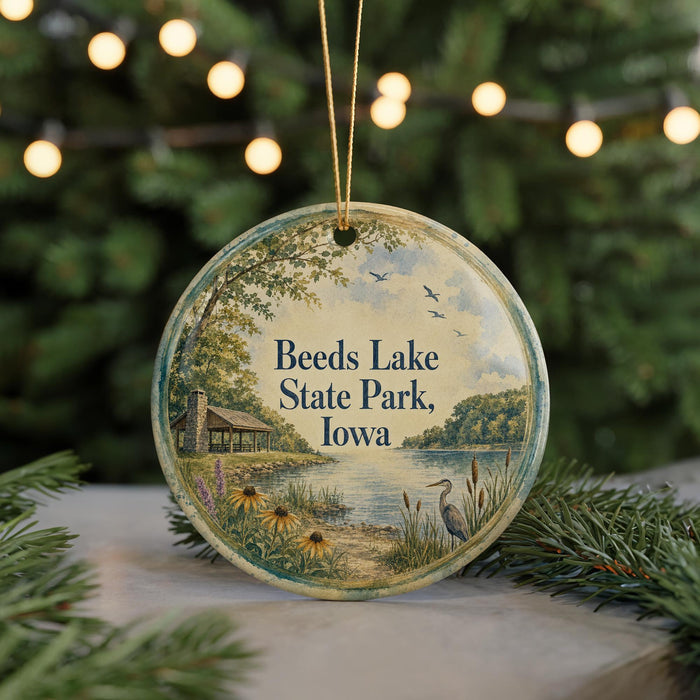 A round ornament with a painting of a lake and a bird, surrounded by greenery, hangs in front of a blurred background with twinkling lights.