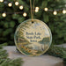 A round ornament with a painting of a lake and a bird, surrounded by greenery, hangs in front of a blurred background with twinkling lights.