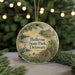 A round ornament with a painting of a house and a bird, surrounded by greenery, hangs in front of a blurred background with twinkling lights.