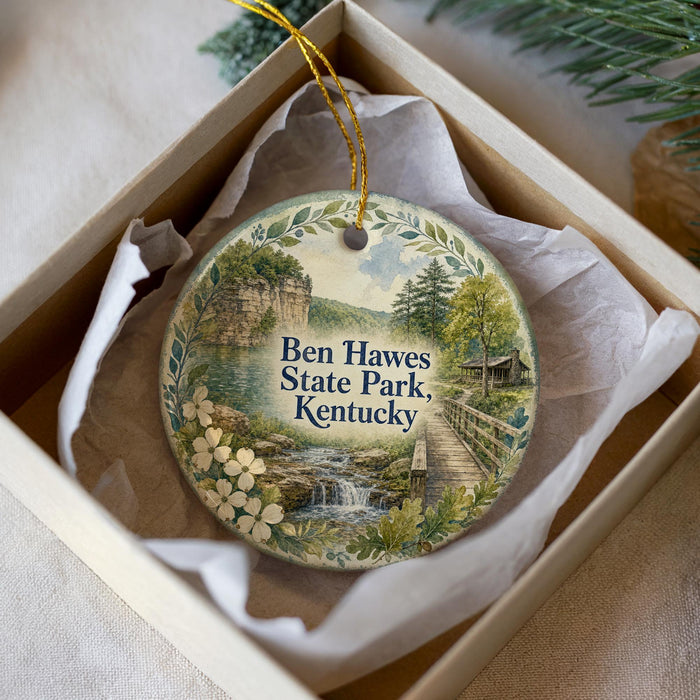 A round ornament with a scenic landscape of Ben Hawes State Park, Kentucky, displayed in a box with a white tissue paper lining.