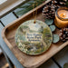 A decorative ornament featuring a scenic landscape of Ben Hawes State Park, Kentucky, with a candle and pine cones on a wooden tray.