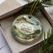 A decorative ornament in a box, featuring a scenic landscape with a lake, mountains, and pine trees.